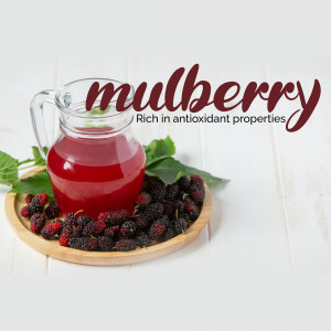 A glass pitcher of mulberry juice with fresh mulberries and leaves on a wooden tray.