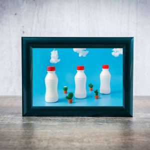 A framed image of three white milk bottles with red caps and small cacti on a blue background.