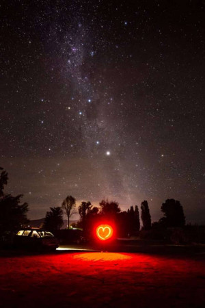 A red heart-shaped light illuminates the foreground under a stunning view of the Milky Way galaxy.