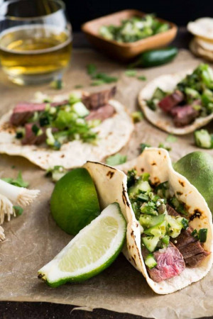 Close-up of steak tacos with vibrant green salsa, lime wedges, and a glass of beverage.