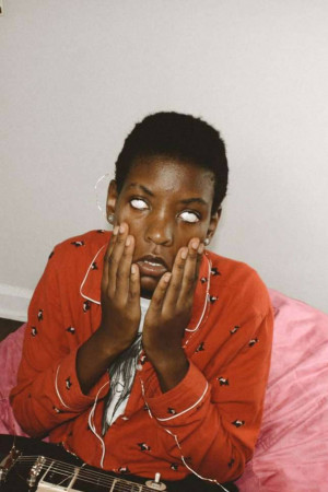 A young Black woman with a surprised expression, holding her face, with a guitar in the background.