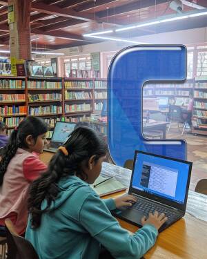 Two young girls using laptops in a library with a large blue 'C' shape in the window.