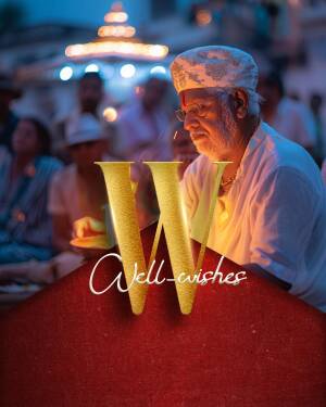 An elderly man in traditional attire holding a lit diya with the text 'Well Wishes' superimposed.
