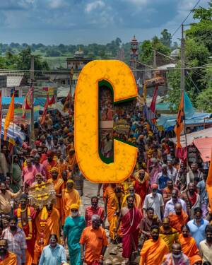 A large crowd of Kanwar pilgrims walking with orange flags and a large letter 'C' superimposed over the scene.