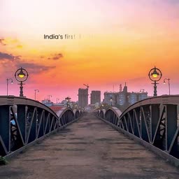 A scenic view looking down the center of an old metallic bridge towards a city skyline during a vibrant pink and orange sunset.