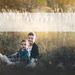 An older brother sitting behind his younger sibling in a sun-drenched grassy meadow at golden hour.
