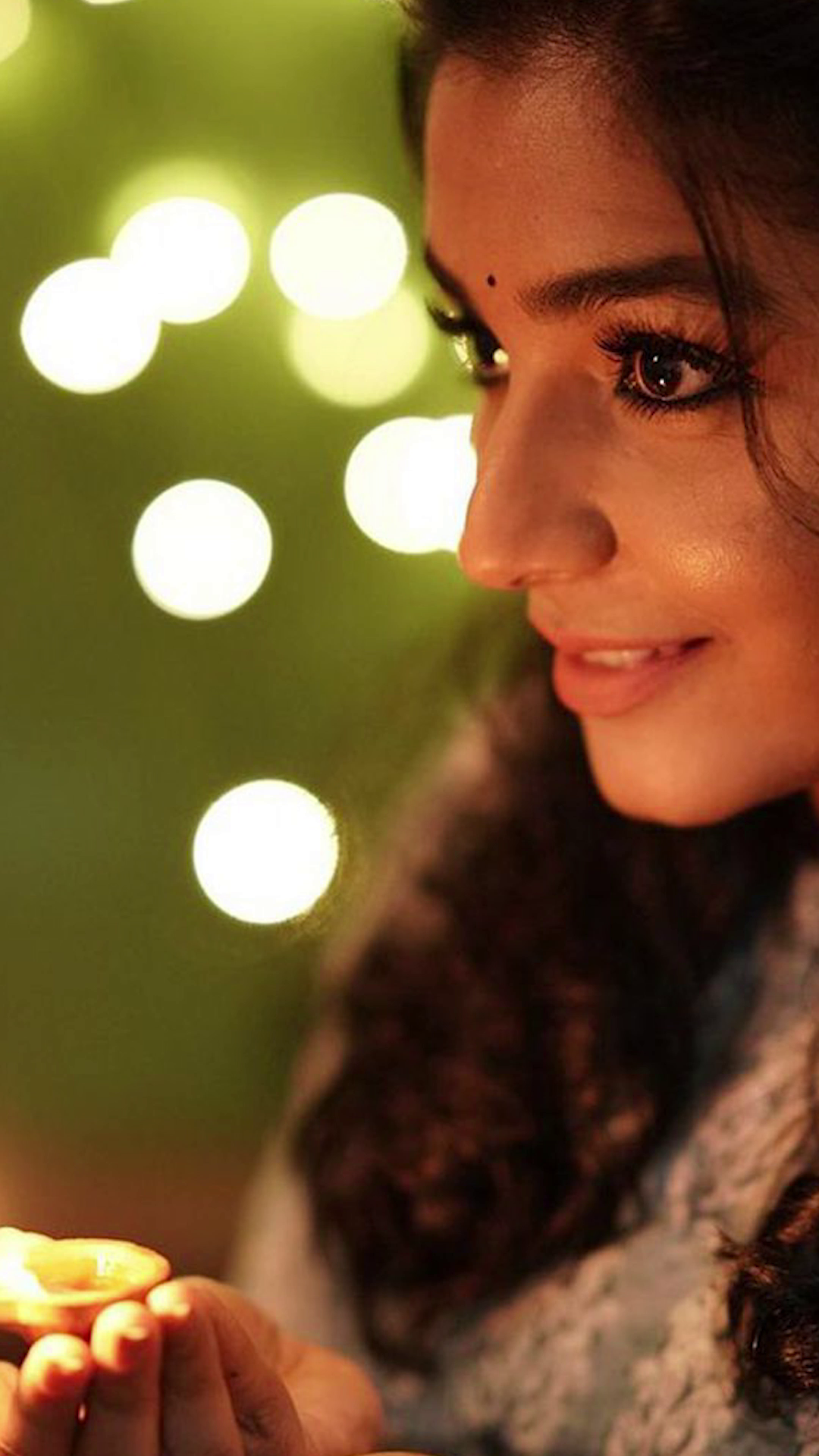 A beautiful Indian woman with a bindi, holding a glowing diya lamp, with bokeh festive lights in the background.