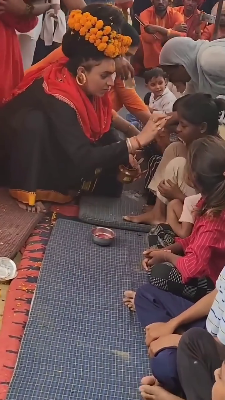 A woman adorned in a black outfit and marigold flower headpiece performing a ritual for a group of children sitting on a mat