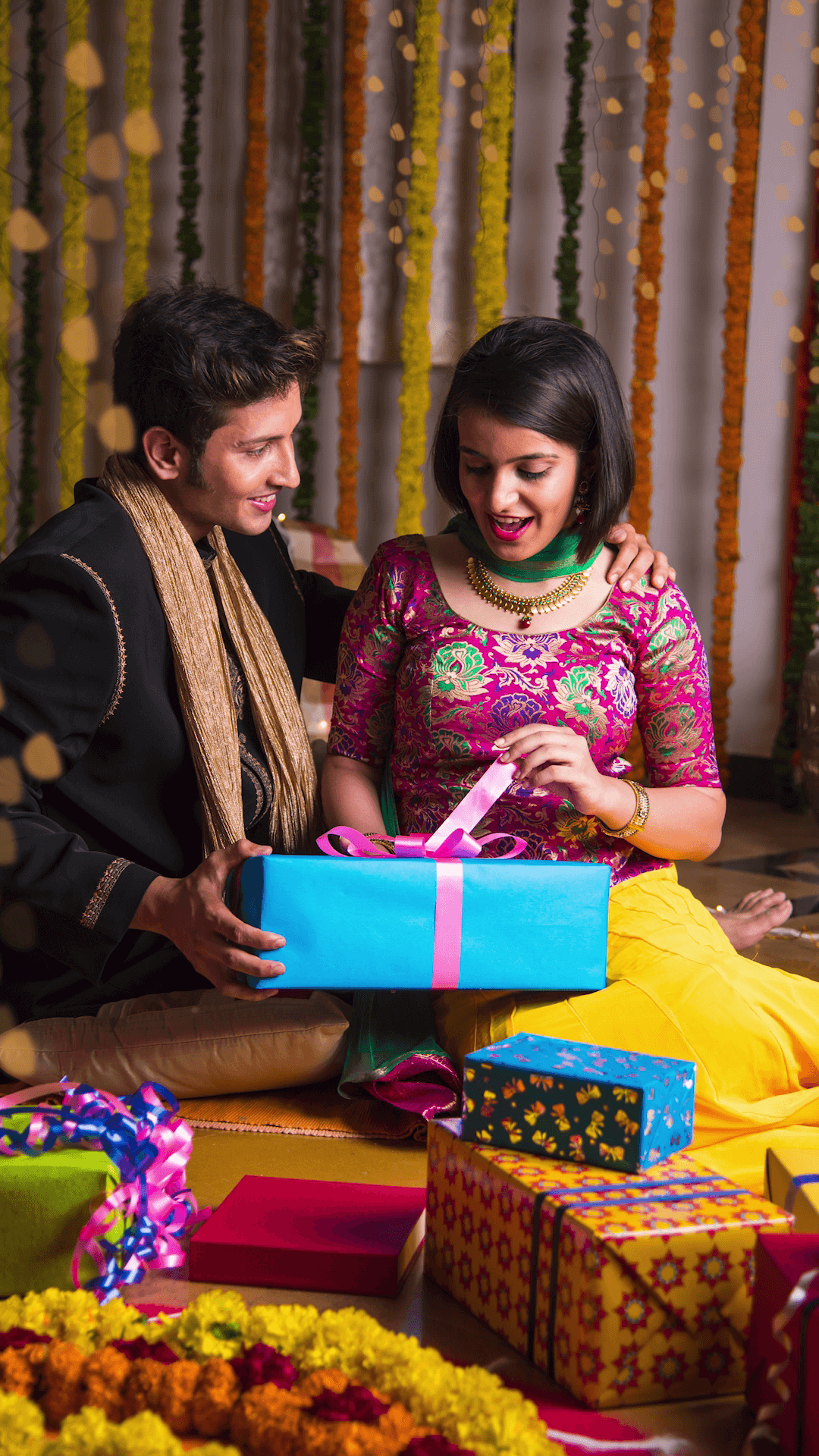 A young Indian couple sitting on the floor surrounded by festive decorations and gift boxes, sharing a joyful moment during Diwali celebrations.