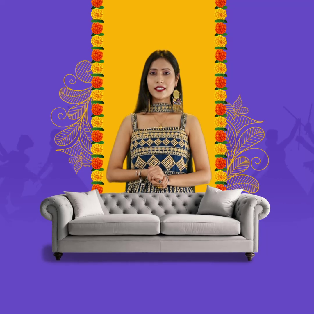 A woman in traditional attire standing behind a grey sofa with a festive yellow and purple background featuring marigold garlands